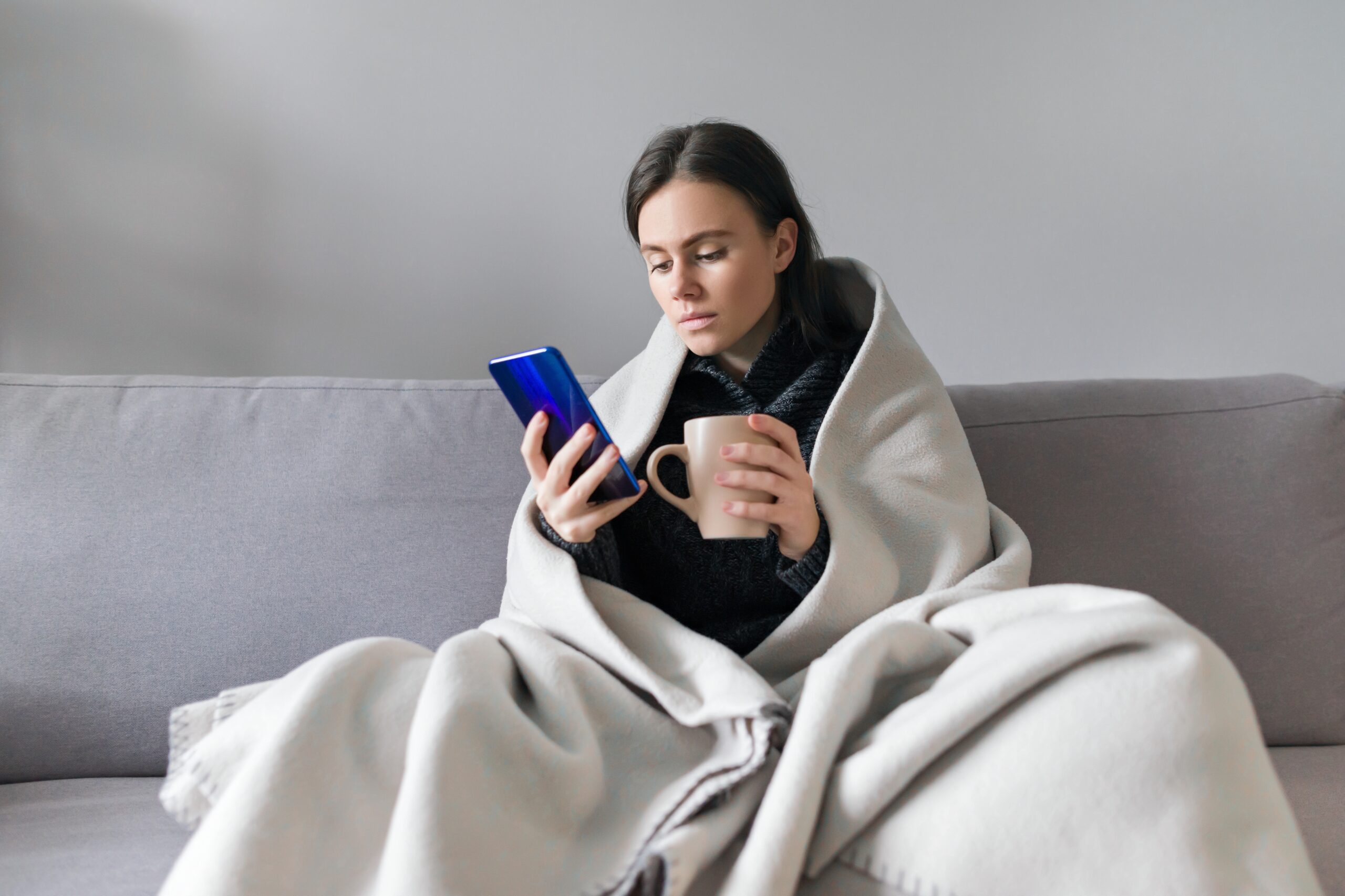 Young woman with cup, smartphone, sitting at home on sofa under warm fleece blanket, copy space